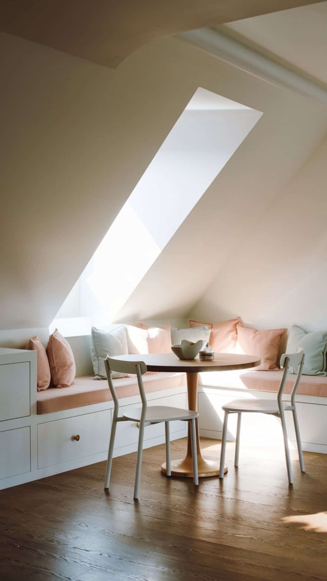 Attic breakfast nook built under sloped ceiling, white built-in bench with hidden storage drawers, soft pastel cushions, small round wooden dining table, slim white chairs, natural light shining through dormer window.